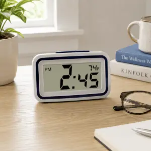 Talking digital clock with temperature display on a desk beside reading glasses, books, and notepad in a bright home setting.